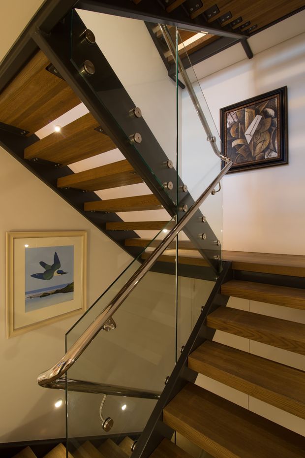 Interior view of a modern multi-level stairway with open-riser timber treads, dark metal stringers and brackets, and clear glass balustrades with polished metal handrails. The stair rises through a tall void, with warm timber ceiling elements, recessed lighting, and framed artwork on the walls.
