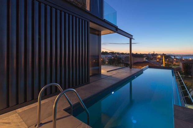 Twilight exterior view of a contemporary elevated terrace with a long narrow swimming pool, dark vertically ribbed facade cladding, slim roof canopy, glass balustrades, and a stainless-steel pool ladder. The terrace overlooks a lit suburban or urban landscape with distant hills and water visible on the horizon.
