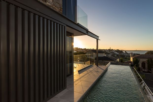 A contemporary multi-level residential exterior at sunset, viewed along a narrow rooftop or elevated terrace beside a long rectangular swimming pool. The facade combines dark vertical ribbed cladding with exposed stonework and extensive glazing, while a glass balustrade frames views over a dense suburban coastal landscape and the water beyond.