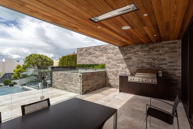A covered outdoor entertaining area with a dark dining table and chairs, built-in stainless-steel barbecue, stone-clad walls and island, tiled flooring, and a warm timber-lined ceiling. Frameless glass balustrades open toward a swimming pool and landscaped residential surroundings.