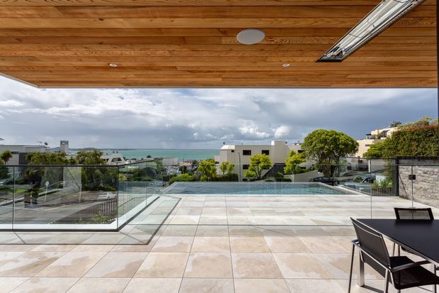 A spacious rooftop or elevated terrace with a large tiled entertaining area, glass balustrades, and a swimming pool overlooking a coastal urban setting. The terrace is partially sheltered by a warm timber-lined ceiling with integrated linear lighting, and includes an outdoor dining table and chairs. Contemporary residential architecture and ocean views create a refined outdoor entertaining environment.