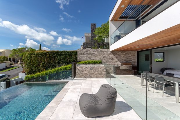A contemporary multi-level residential exterior viewed from a pool terrace. The scene features a rectangular swimming pool, pale large-format tiled paving, glass balustrades, a grey stone-clad retaining wall and stair structure, and a projecting upper level with white rendered walls and warm timber soffits. Outdoor dining furniture and a beanbag sit beside the pool, with suburban homes and landscaped hedges beyond.
