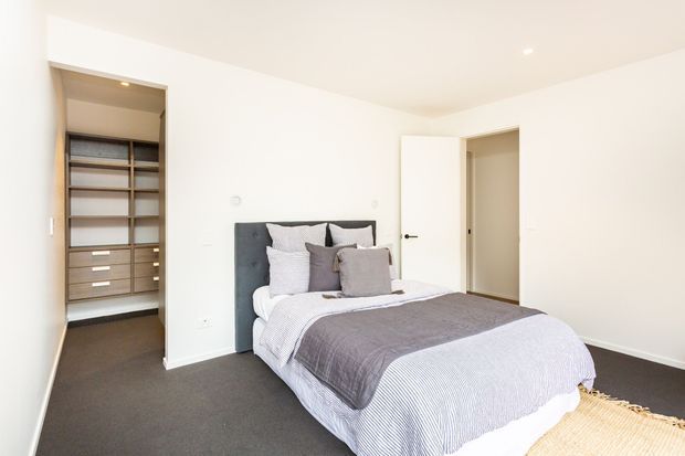 Bright, modern bedroom with a neatly made bed, upholstered charcoal headboard, layered grey and white bedding, dark carpet, and a walk-in wardrobe visible through an open doorway. White walls, recessed lighting, and a simple ensuite-style or hallway door contribute to a clean, understated interior.