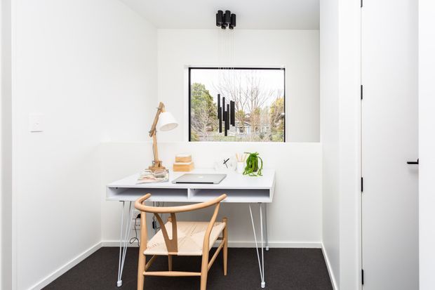 Bright compact home office with white walls and built-in white desk beneath a black-framed window. A light timber wishbone-style chair, desk lamp, laptop, books and small plant furnish the workspace. The room has dark carpet flooring, minimal black ceiling lighting and a white door.
