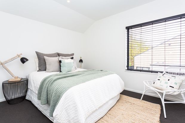 Bright, compact bedroom with white walls and ceiling, a neatly made bed layered with white, grey, and pale sage-green bedding, a black wire side table, articulated black desk lamp, and a white lounge chair beside a large window with dark horizontal blinds. A light woven rug adds texture over the dark floor.