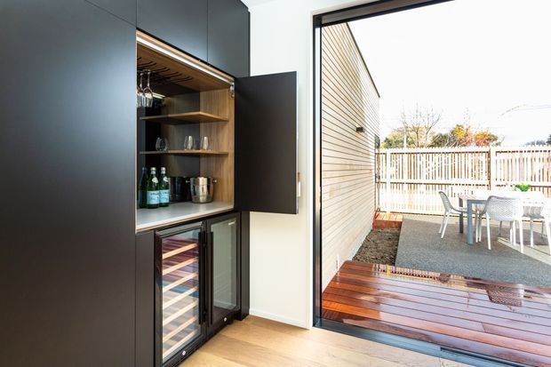 A compact built-in drinks bar and refrigerated wine storage unit in a dark matte joinery wall, with open timber shelving and glassware. Large sliding or folding glazing opens directly onto a timber deck and paved outdoor dining area with a white fence beyond.