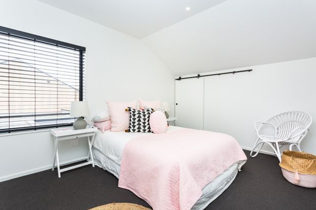 Bright, compact bedroom with white walls and ceiling, dark carpet, a large window with black horizontal blinds, and a white bed dressed in pale pink bedding and decorative cushions. A small white bedside table, white wire lounge chair, woven basket, and black barn-style sliding door hardware add simple contemporary character.