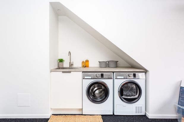 Compact modern laundry tucked beneath a sloping ceiling, with white built-in cabinetry, a grey stone-look benchtop, undermount sink and tap, and two front-loading white appliances. The space has minimal styling and bright neutral finishes.