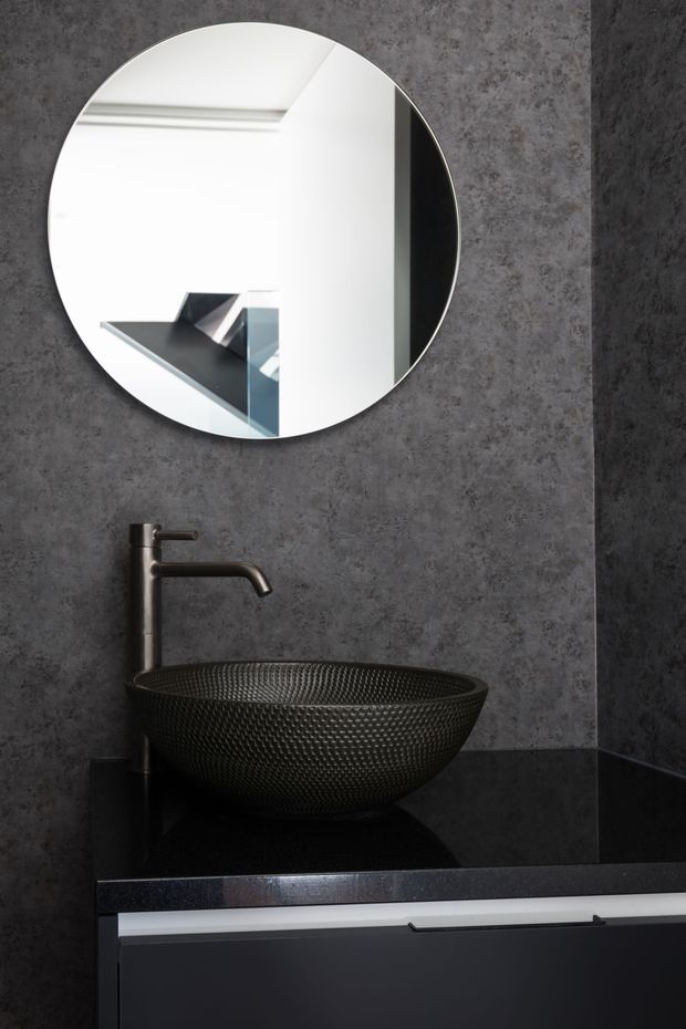A compact, dark contemporary bathroom vanity featuring a textured black vessel basin, matte dark metal wall-mounted tap, black polished benchtop, and dark grey concrete-look wall finish. A large round frameless mirror reflects a bright adjacent interior with angular architectural forms. The lower vanity has dark cabinetry with a slim contrasting light drawer front.