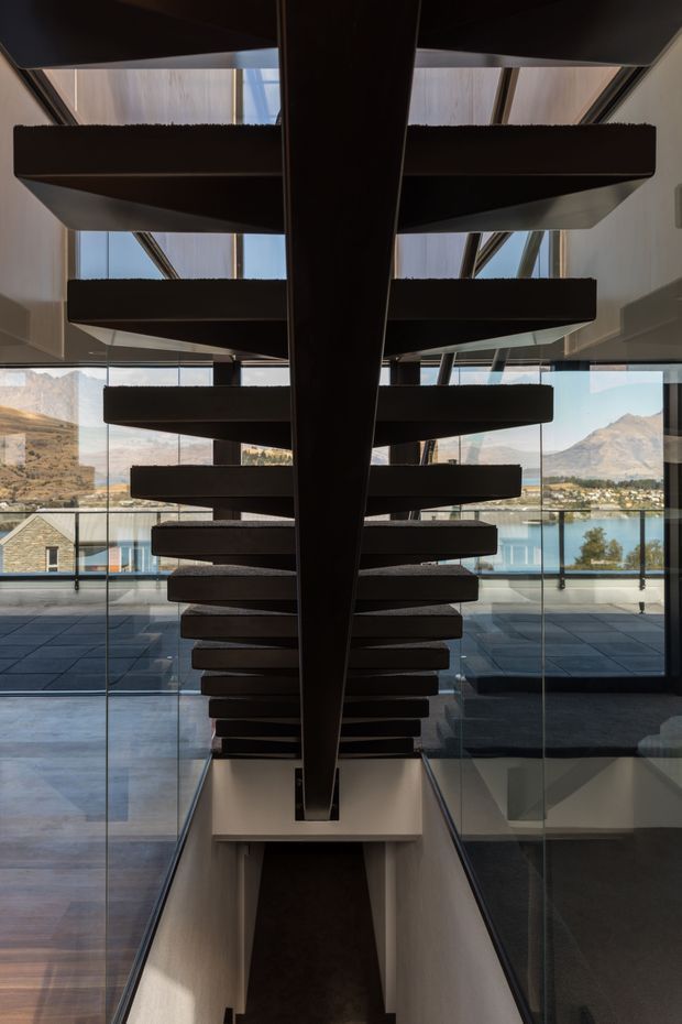 Interior architectural photograph looking along a dramatic dark timber or metal open-riser staircase with a central structural spine and cantilevered treads. Full-height glazing and glass balustrades surround the stair, opening views toward a lake and mountainous landscape. The space is contemporary, highly geometric, and light-filled, with dark stair elements contrasting against pale walls and blue-grey flooring.
