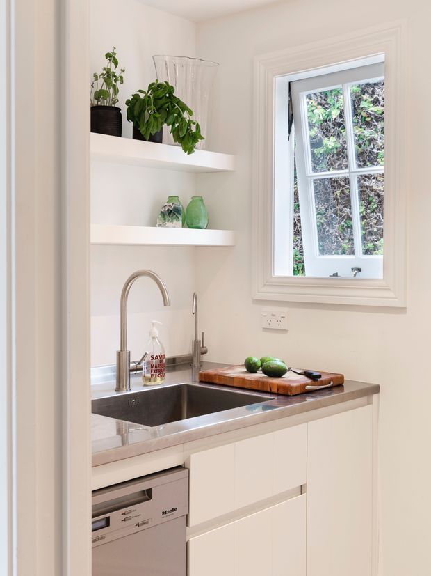 Bright compact kitchen or scullery with white built-in cabinetry, open shelving, stainless-steel sink and dishwasher, and a timber chopping board on the benchtop. A traditional multi-pane window with white trim provides natural light, while potted plants and green glassware add an organic accent.