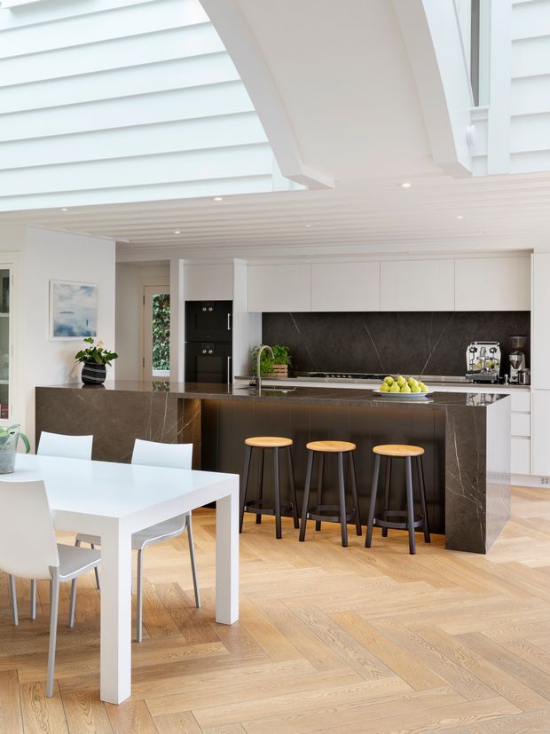 Bright open-plan kitchen and dining area with white cabinetry, a dark stone island and matching backsplash, timber herringbone flooring, and a white dining table with contemporary chairs. Three timber-seat bar stools line the island. The room features a white slatted ceiling with a large skylight or clerestory opening and abundant natural light.