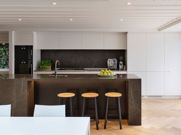 A refined contemporary kitchen with a large dark stone island and integrated breakfast bar, three timber-topped stools, white full-height cabinetry, dark stone splashback, integrated black appliances, and a pale herringbone timber floor. A dining table is partially visible in the foreground. The ceiling features painted linear timber or panel detailing with recessed lighting.