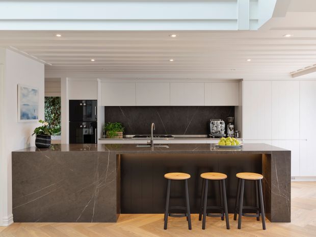 Bright contemporary kitchen with a large dark stone island and integrated breakfast bar, three timber-and-black stools, white flat-panel cabinetry, dark stone splashback, and a white slatted timber ceiling with recessed lighting. Light herringbone timber flooring and minimal styling create a refined, high-contrast interior.