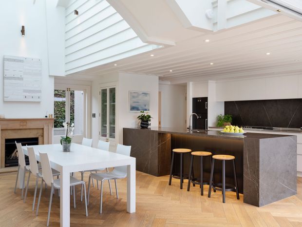 Bright, open-plan kitchen and dining area with a large dark stone island and breakfast bar, white flat-panel cabinetry, dark stone splashback, and a white dining table with contemporary chairs. The room features a high slatted ceiling with skylight-like glazing, recessed lighting, pale herringbone timber flooring, and generous glazed doors opening toward greenery. A fireplace is visible at the edge of the dining area.