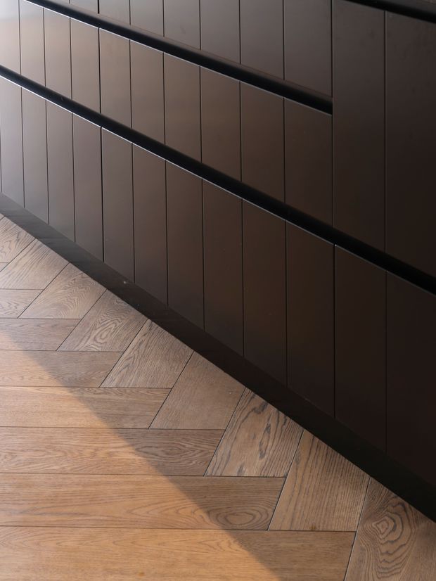 Close-up architectural interior detail showing a dark, vertically panelled built-in wall or cabinetry with black horizontal trims, beside a warm natural timber herringbone/parquet floor. Strong diagonal composition and soft directional light create a refined, moody atmosphere.