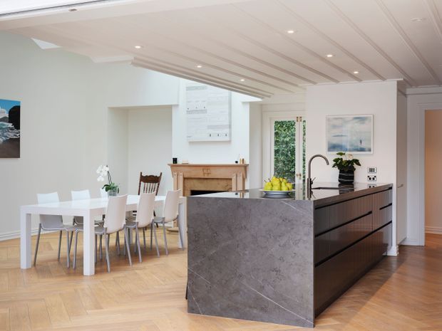 Bright open-plan kitchen and dining area with a large dark stone island, integrated sink and gooseneck tap, white dining table and chairs, timber herringbone flooring, a traditional fireplace, and a white painted ceiling with linear recessed detailing. The space combines contemporary kitchen joinery with traditional architectural elements.