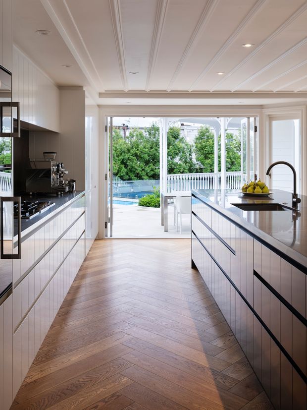 Bright, refined kitchen viewed along a central galley layout. White paneled cabinetry lines both sides, with dark benchtops, integrated appliances and a prominent curved gooseneck tap. The room opens through wide glazed doors to a veranda and swimming pool, while the ceiling features detailed linear panel moulding and recessed downlights. Warm timber herringbone flooring provides contrast.