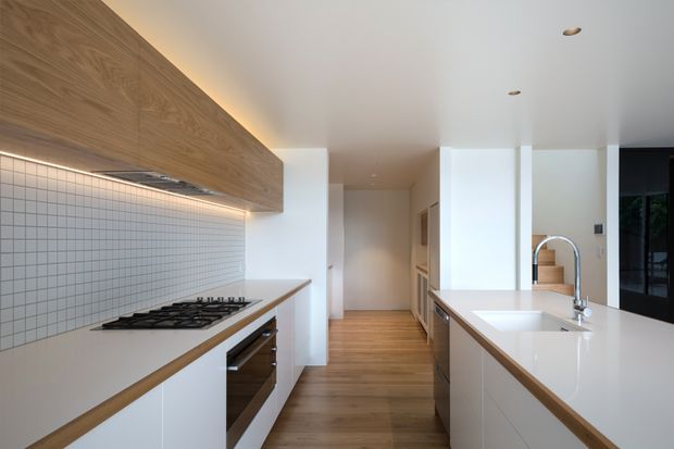 A bright contemporary galley kitchen with white cabinetry, white benchtops, a tiled grid-pattern splashback, integrated gas cooktop and oven, timber flooring, and a large timber-faced overhead feature cabinet with concealed lighting. A central island with an undermount sink and curved chrome tap runs along the right side, with a hallway and dark-framed glazed door visible beyond.