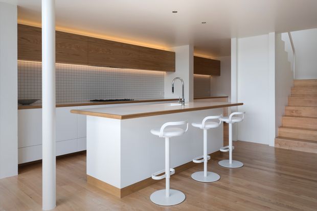 Bright open-plan kitchen with a large white island, timber benchtop, white bar stools, warm timber cabinetry, tiled splashback, and timber flooring. A timber staircase is visible at the right, with concealed warm LED lighting highlighting the upper cabinetry.