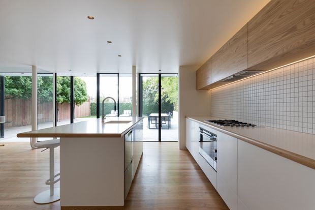 Contemporary open-plan kitchen with a large central island, white cabinetry, integrated appliances, timber flooring, a white square-tile splashback, and extensive floor-to-ceiling glazing opening to an outdoor area. Warm timber overhead cabinetry and under-cabinet lighting add contrast to the restrained white fit-out.