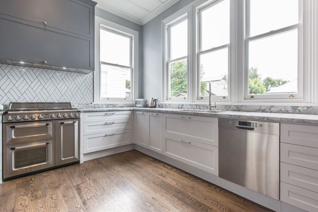 Bright, spacious kitchen with grey shaker-style cabinetry, white herringbone tile backsplash, marble-look countertops, stainless-steel range and dishwasher, and warm timber flooring. Large traditional sash windows provide abundant natural light and views of neighbouring homes and greenery.