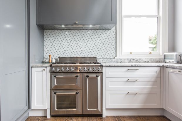 A bright, compact contemporary kitchen featuring a stainless-steel range cooker beneath a dark grey rangehood, white shaker-style cabinetry, a white herringbone tile splashback, pale stone-look benchtops, and timber flooring. A window and small countertop appliances are visible on the right.