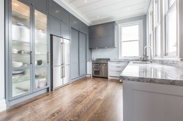 Bright, refined kitchen with extensive blue-grey built-in cabinetry, glass-front display doors, stainless-steel appliances, a marble-patterned benchtop and splashback, warm timber flooring, and a white painted tongue-and-groove ceiling. Large windows provide abundant natural light.
