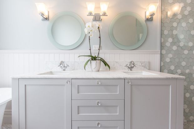 Bright, refined bathroom vanity featuring a pale painted double vanity with recessed-panel cabinetry, white marble benchtop and backsplash, twin round mirrors, decorative wall sconces, white beadboard wainscoting, and a grey hexagonal tile feature wall. A small orchid adds a soft natural accent.