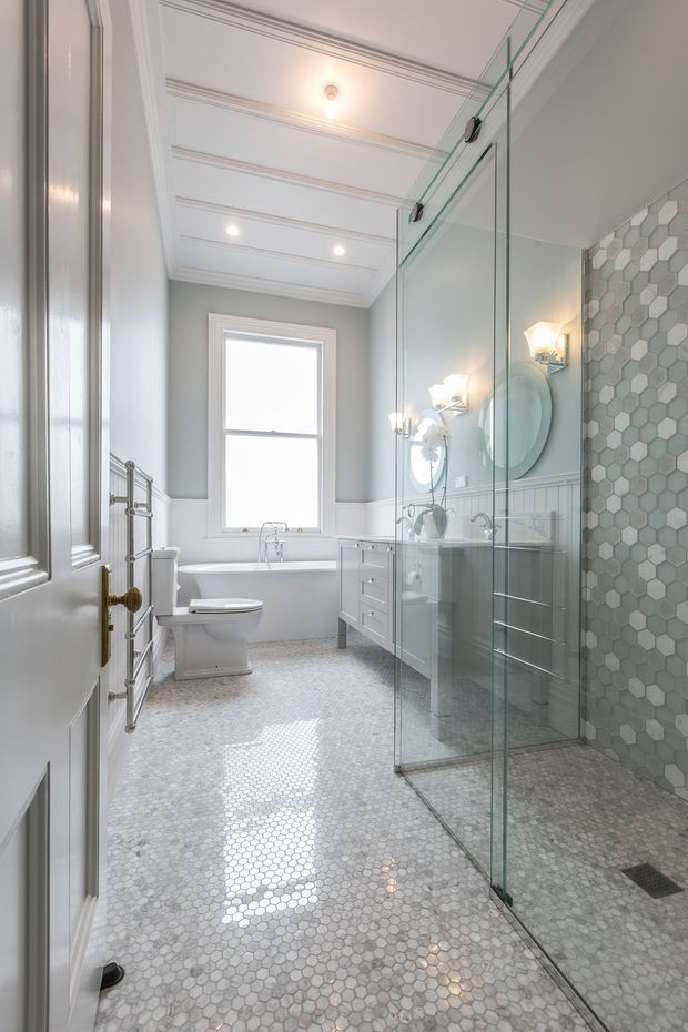 Bright, refined bathroom featuring a glass-enclosed walk-in shower, freestanding bathtub beneath a tall window, toilet, pale vanity, hexagonal mosaic floor and shower-wall tiles, grey painted walls with white wainscoting, ornate white coffered ceiling, and traditional metal fixtures.