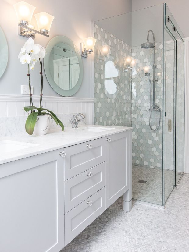 Bright, refined bathroom featuring a pale grey vanity with shaker-style cabinetry, a white stone benchtop, chrome tapware, mirrored medicine cabinet, glass-enclosed walk-in shower, and hexagonal mosaic wall and floor tiles. Soft globe-style wall sconces, white beadboard wall panelling, and an orchid add a classic yet fresh decorative character.