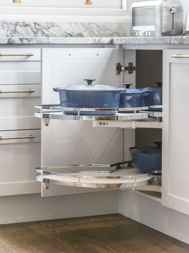 Close-up of a white kitchen corner cabinet with two pull-out rotating shelves holding blue cookware. The kitchen features white cabinetry with long metallic handles, a marble-patterned benchtop and backsplash, stainless or chrome shelf hardware, and dark timber flooring. A toaster and rounded appliance sit on the benchtop.
