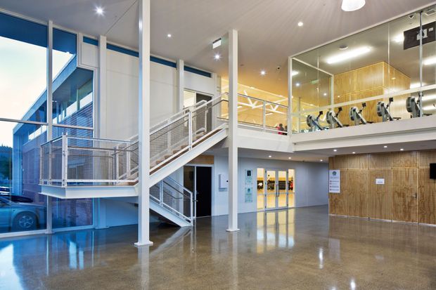 A bright contemporary multi-level commercial interior with a polished concrete floor, exposed structural columns, a central metal staircase and mezzanine walkway, extensive glazing, and a reception or circulation area. A gym or fitness area with exercise equipment is visible behind glass on the upper level. The space has a clean institutional or commercial character with white walls, timber-toned fixed wall panelling, and abundant daylight.