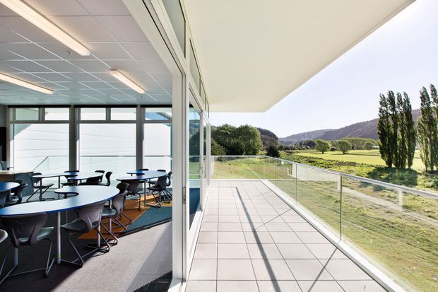 A modern commercial building with a glazed meeting or collaboration room opening onto a wide tiled terrace. The interior contains multiple round tables and office chairs beneath a suspended grid ceiling, while the terrace has frameless glass balustrading and overlooks a landscaped rural valley with hills beyond.