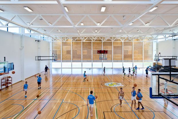 A bright, expansive indoor basketball gymnasium with a polished timber court marked for multiple sports, several basketball hoops, players practising and playing, white-painted exposed structural trusses, high ceilings, clerestory-style glazing, and a timber-panelled feature wall at the far end.