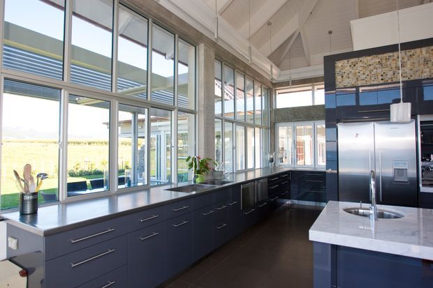 A spacious contemporary kitchen with extensive floor-to-ceiling glazing, dark navy built-in cabinetry, long dark benchtops, and a central island with a pale stone benchtop and integrated sink. The room has a high pitched white ceiling with exposed structural beams, polished concrete-style columns, pendant lights, and a mosaic-tile feature strip above the cabinetry. Rural landscape is visible beyond the windows.