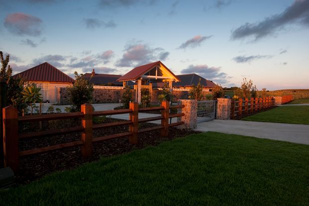 A single-storey rural-style property viewed from a landscaped exterior at dusk. The complex has multiple pitched and gabled roofs, warm timber framing, stone masonry walls, metal roofing, and a covered central entry or pavilion. Timber post-and-rail fencing, a paved driveway or walkway, lawn, and open agricultural land frame the building.