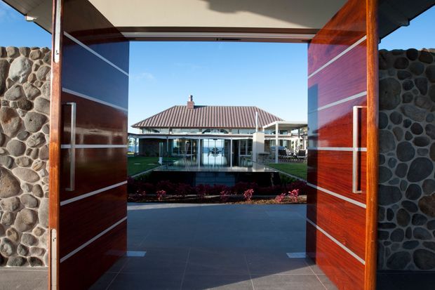 A dramatic contemporary entryway viewed through wide open timber pivot doors. The doors feature rich reddish-brown wood panels with horizontal metal inlays and long vertical handles, set within walls clad in rounded dark stone. Beyond the threshold is a landscaped courtyard with a reflective rectangular water feature and a glazed pavilion or living area, with a low-pitched tiled roof visible in the background.