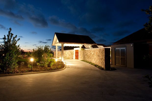 A dusk exterior view of a landscaped residential property, showing a broad illuminated concrete driveway curving past planted garden beds toward a detached-looking gabled structure. A rough light stone or masonry boundary wall, timber door, pitched dark roof, and warm exterior lighting define the scene. The image is primarily an approach and driveway view, with no interior spaces visible.