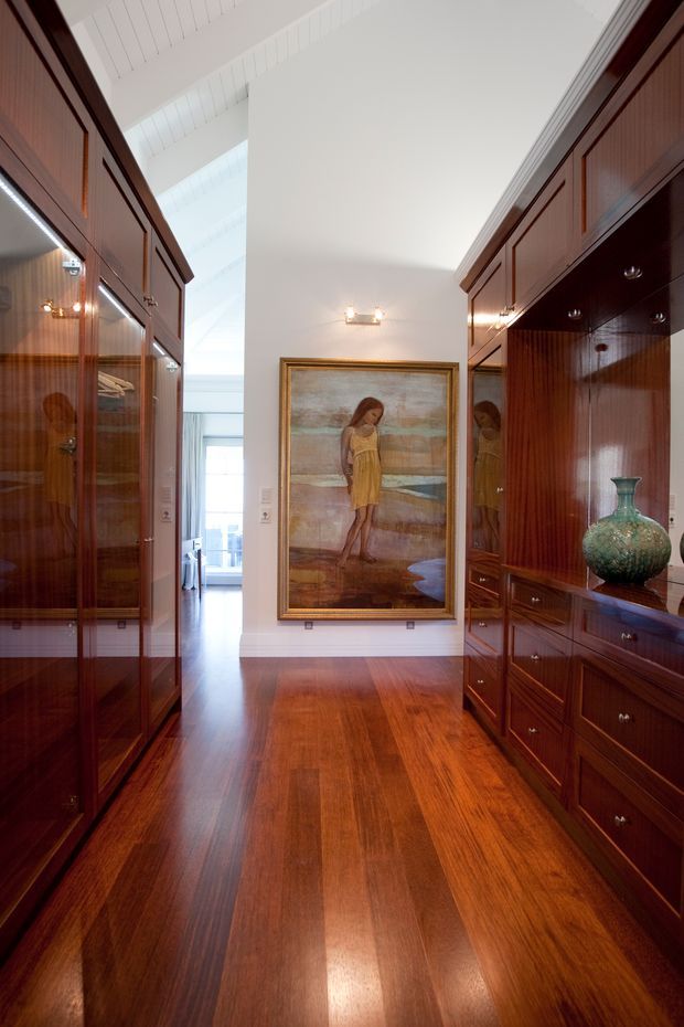 A refined, narrow interior corridor or gallery lined on both sides with extensive dark timber built-in cabinetry and glazed display cabinets. The polished timber floor matches the rich cabinetry, while a tall white vaulted or pitched ceiling and clerestory-like glazing bring in natural light. A framed painting forms the focal point at the end of the passage, with integrated cabinet lighting and a decorative ceramic vase adding detail.