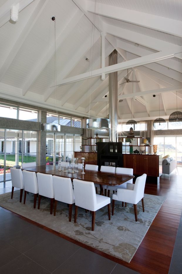Bright, spacious dining area in a high-ceilinged open-plan interior. A large dark timber dining table is surrounded by white upholstered dining chairs on a patterned rug. Behind it is a dark timber sideboard and shelving with a black fireplace or stove, while extensive glazing provides views to an outdoor courtyard. The room features prominent white-painted timber cathedral ceilings, exposed white beams and rafters, suspended woven pendant lights, polished timber flooring, and dark tiled flooring at the foreground.