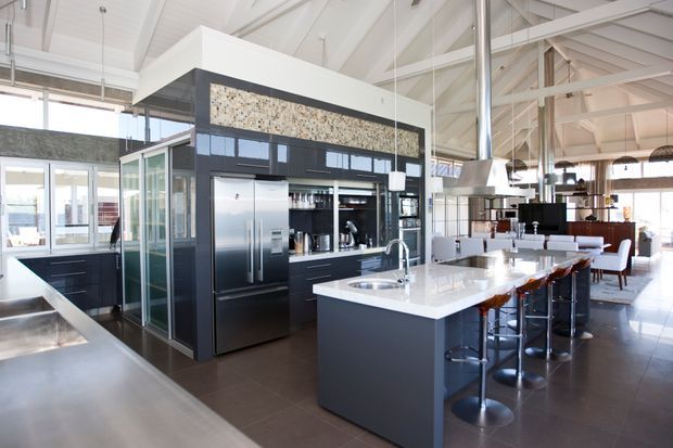 A spacious open-plan kitchen in a bright contemporary interior, featuring a large dark island with a white stone benchtop and integrated sink, dark glossy cabinetry, stainless-steel appliances, and bar seating. A distinctive terrazzo-like feature strip sits above the cabinetry. The kitchen opens to a dining and living area, with exposed white roof trusses, high ceilings, extensive glazing, and a large stainless-steel rangehood creating an airy industrial-contemporary character.