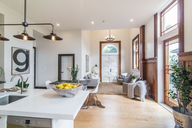 Bright open-plan interior combining a contemporary kitchen with a welcoming entry and living area. The room features a large white island, pale timber flooring, white walls, dark timber window and door frames, tall glazing, pendant lighting, and a paneled front door. A dining table and seating area sit near the entry, with decorative plants and artwork adding warmth.