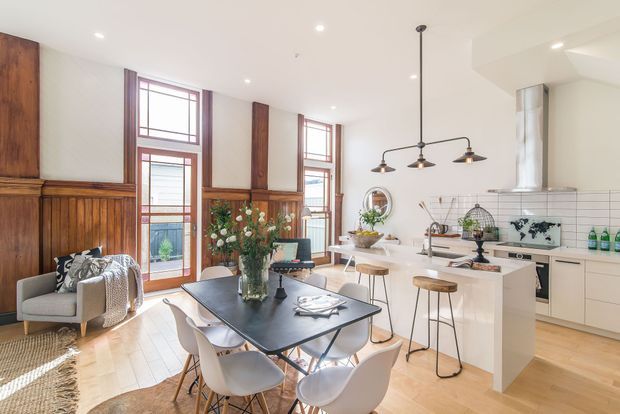 Open-plan kitchen and dining area in a bright converted heritage-style interior, featuring warm timber wall panelling, tall divided windows and doors, pale timber flooring, a white kitchen with island, black-framed dining table, pendant lighting, and contemporary dining chairs. The space blends traditional architectural detailing with modern furnishings and finishes.