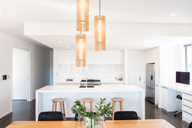Bright contemporary open-plan kitchen and dining area with a large white island, timber bar stools, black dining chairs, white cabinetry, integrated stainless-steel appliances, dark timber flooring, and three vertical timber pendant lights.