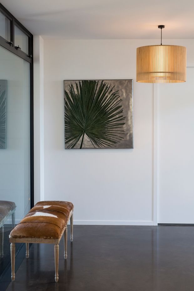 A refined minimalist interior vignette with white paneled walls, dark polished flooring, black-framed glazing, and a pair of brown leather upholstered benches. A large tropical leaf artwork and a warm woven pendant light add natural texture and contrast.