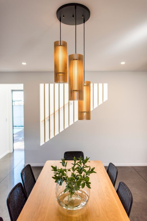 Bright contemporary dining room with a long light timber dining table, dark woven dining chairs, and a clear glass vase of greenery. Three suspended cylindrical pendant lights with warm timber slat shades form the main feature, while a glass-panel stair balustrade is visible in the background. The room has white walls, recessed ceiling lights, and a polished concrete floor.