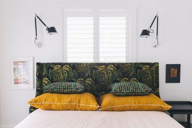 A bright, compact bedroom with a white bed dressed in mustard-gold and patterned cushions, backed by a dark tropical botanical upholstered headboard. White plantation shutters sit between two black wall-mounted swing-arm lamps, with framed artwork and a slim dark console completing the room.