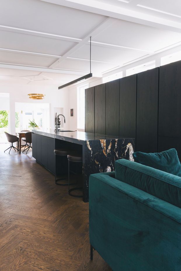 Bright open-plan kitchen and dining area with a dark, full-height kitchen wall and island, a dramatic black stone island end, rich herringbone timber flooring, teal upholstered seating, and contemporary pendant lighting. White coffered-style ceiling detailing and large windows create an airy contrast to the dark joinery.