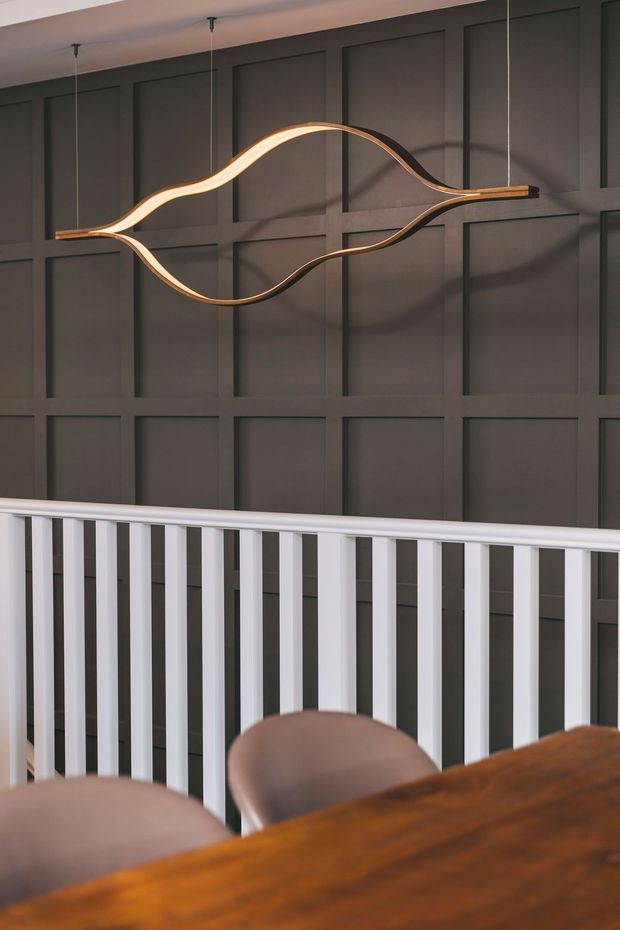 Interior view of a refined dining or landing area featuring a dark charcoal wall with geometric recessed panel moulding, a sculptural suspended wave-shaped pendant light with warm illumination, and a white vertical balustrade. The foreground includes a warm timber dining table and upholstered dining chairs.